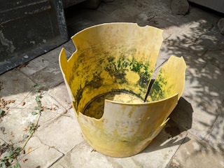 A broken yellow plastic container lies on a stone pavement. It has jagged edges and green stains inside, suggesting wear and exposure to the elements. Dried leaves and scattered twigs are visible around the container, emphasizing a neglected or abandoned environment.