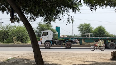 A flatbed truck with a long trailer is parked on a road beside some greenery, while a man rides a motorcycle alongside. Trees and electrical wires in the background add to the rural setting.
