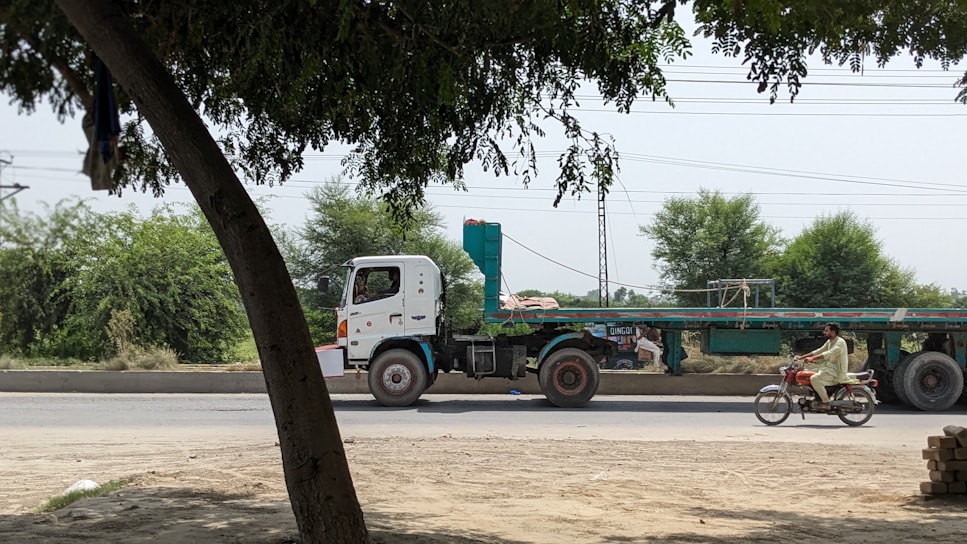 A flatbed truck with a long trailer is parked on a road beside some greenery, while a man rides a motorcycle alongside. Trees and electrical wires in the background add to the rural setting.