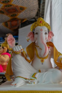 A detailed statue of a deity with an elephant head, wearing elaborate golden headgear and jewelry. The deity is seated in a cross-legged position, one hand raised in a blessing gesture. There are decorative patterns on the background fabric.