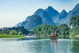 A serene river scene with lush green vegetation and towering karst mountains in the background. A group of people wearing life jackets are standing on a bamboo raft, being guided by someone with a pole. The water reflects the greenery and the sky, adding to the tranquil ambiance. Nearby, more rafts can be seen, likely part of a larger group or tour.