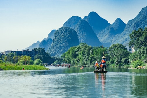 A serene river scene with lush green vegetation and towering karst mountains in the background. A group of people wearing life jackets are standing on a bamboo raft, being guided by someone with a pole. The water reflects the greenery and the sky, adding to the tranquil ambiance. Nearby, more rafts can be seen, likely part of a larger group or tour.