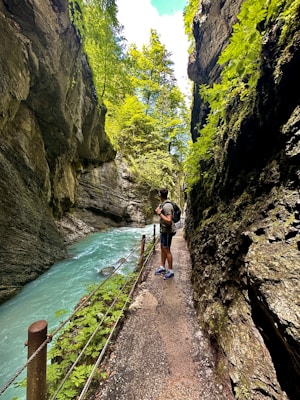 A narrow path runs alongside a turquoise river, bordered by high rocky cliffs covered with greenery. A person wearing a backpack stands observing the scenery, surrounded by lush vegetation and vibrant, overhanging trees.