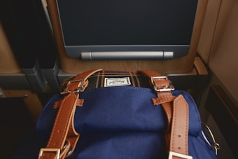 A blue fabric bag with brown leather straps is placed in front of an airplane or train seat equipped with a screen. The seat is made of a combination of materials including wood and plastic.