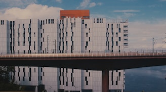 A modern architectural building with a geometric facade, featuring white panels and dark windows. The structure is positioned behind an elevated bridge, and a portion of bright orange building is visible at the top. The sky is partly cloudy, and there is a large body of water in the background.