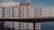 A modern architectural building with a geometric facade, featuring white panels and dark windows. The structure is positioned behind an elevated bridge, and a portion of bright orange building is visible at the top. The sky is partly cloudy, and there is a large body of water in the background.