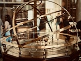 A brass armillary sphere is displayed prominently, featuring intricate rings and inscriptions depicting astronomical concepts. Several people in the background suggest a museum or educational setting.