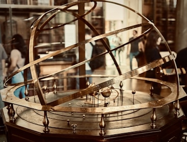 A brass armillary sphere is displayed prominently, featuring intricate rings and inscriptions depicting astronomical concepts. Several people in the background suggest a museum or educational setting.