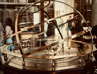 A brass armillary sphere is displayed prominently, featuring intricate rings and inscriptions depicting astronomical concepts. Several people in the background suggest a museum or educational setting.