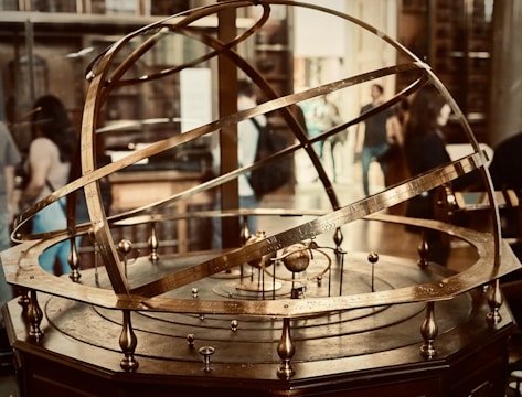 A brass armillary sphere is displayed prominently, featuring intricate rings and inscriptions depicting astronomical concepts. Several people in the background suggest a museum or educational setting.
