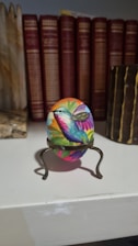 A beautifully painted egg featuring a vibrant depiction of a hummingbird perched on a small metal stand. The surrounding background consists of an organized row of books with red and brown spines as well as natural stone elements.