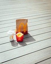 A paper bag, fries container, and drink cup are placed on a light-colored wooden deck. The items prominently display the McDonald's logo in yellow on a red background.