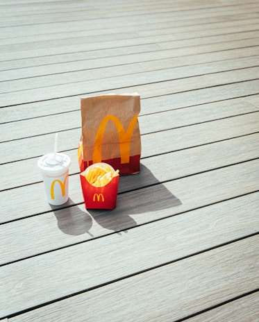 A paper bag, fries container, and drink cup are placed on a light-colored wooden deck. The items prominently display the McDonald's logo in yellow on a red background.