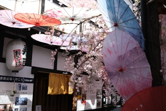The image features a decorative setup with cherry blossom branches and colorful traditional paper umbrellas hanging from the ceiling. A Japanese lantern with text and some posters are visible on the wall in the background, along with a rustic wooden door partially covered by a yellow curtain.