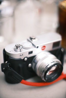 A vintage-style camera with a silver finish and black body, featuring a large lens and a red strap attached. The camera has a classic design with visible dials and a textured grip.