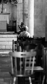 A black and white image of the interior of a church, featuring a solitary person sitting on a chair near an altar. The setting includes stone walls and rows of empty chairs, contributing to a serene and contemplative atmosphere.