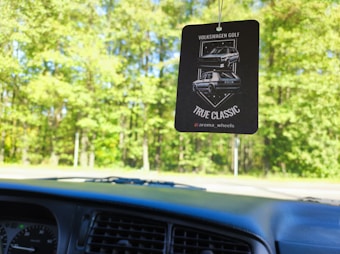 A car air freshener hangs from the rearview mirror, depicting a 'Volkswagen Golf' with the words 'True Classic'. The interior of the car is partially visible in the foreground, while a lush, green forest with sunlight filtering through the leaves forms the background.