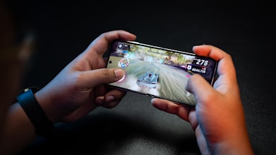 Hands holding a smartphone with a racing game on the screen, featuring a car speeding along a track with visible game controls and speed indicators.