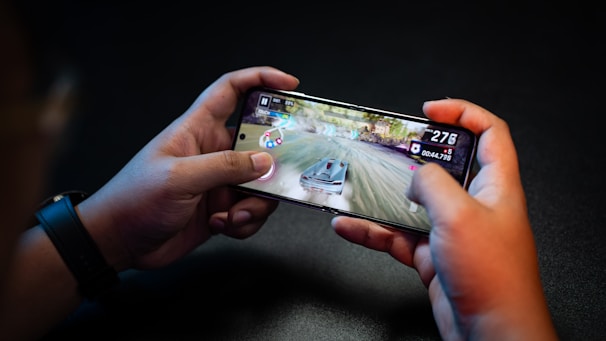 Hands holding a smartphone with a racing game on the screen, featuring a car speeding along a track with visible game controls and speed indicators.