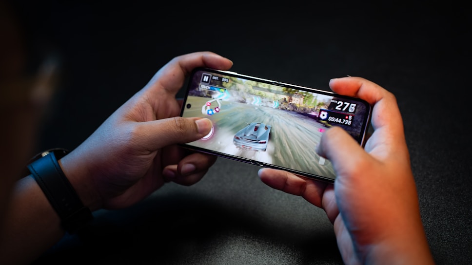 Hands holding a smartphone with a racing game on the screen, featuring a car speeding along a track with visible game controls and speed indicators.