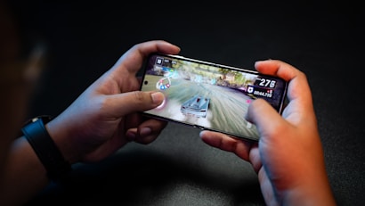 Hands holding a smartphone with a racing game on the screen, featuring a car speeding along a track with visible game controls and speed indicators.