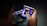 A person is holding a small electronic device, possibly a smartphone with a foldable screen, playing a video game. The game features a dark, fiery theme with icons or characters displayed on the screen.