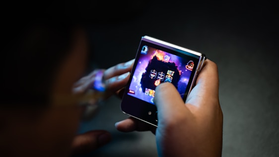 A person is holding a small electronic device, possibly a smartphone with a foldable screen, playing a video game. The game features a dark, fiery theme with icons or characters displayed on the screen.
