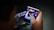 A person is holding a small electronic device, possibly a smartphone with a foldable screen, playing a video game. The game features a dark, fiery theme with icons or characters displayed on the screen.
