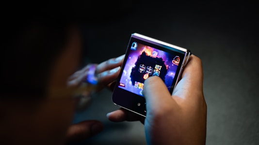 A person is holding a small electronic device, possibly a smartphone with a foldable screen, playing a video game. The game features a dark, fiery theme with icons or characters displayed on the screen.