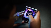 A person is holding a small electronic device, possibly a smartphone with a foldable screen, playing a video game. The game features a dark, fiery theme with icons or characters displayed on the screen.