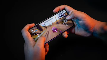 Hands holding a smartphone with a racing game displayed on the screen. The image shows a vibrant racing scene with a red and white car moving at high speed along a desert-like track. Various on-screen controls and speed indicators are visible.