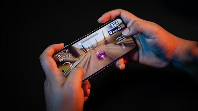 Hands holding a smartphone with a racing game displayed on the screen. The image shows a vibrant racing scene with a red and white car moving at high speed along a desert-like track. Various on-screen controls and speed indicators are visible.