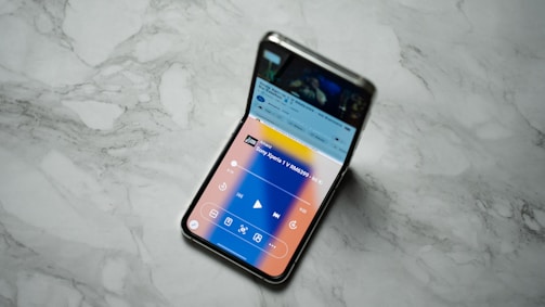 A foldable smartphone is placed on a marble surface with its screen partially folded. The screen displays a user interface with media controls and a notification bar, indicating the phone is in use.