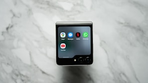 A foldable smartphone is placed on a marble surface with its external screen displaying several app icons, including Maps, Messages, Netflix, WhatsApp, and YouTube.