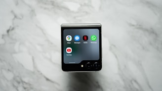 A foldable smartphone is placed on a marble surface with its external screen displaying several app icons, including Maps, Messages, Netflix, WhatsApp, and YouTube.