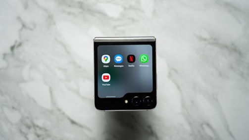 A foldable smartphone is placed on a marble surface with its external screen displaying several app icons, including Maps, Messages, Netflix, WhatsApp, and YouTube.
