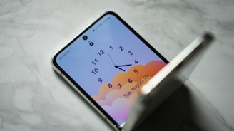 A smartphone with a foldable screen is partially open on a marble surface. The screen displays an analog clock with numbers 1 through 12 and hands pointing to around 4:30. The background of the screen features a gradient of soft orange and pink colors, resembling clouds. The date displayed on the screen is Tuesday, August 15.