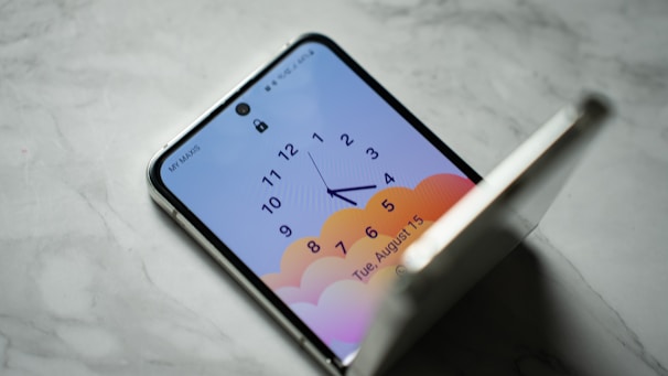A smartphone with a foldable screen is partially open on a marble surface. The screen displays an analog clock with numbers 1 through 12 and hands pointing to around 4:30. The background of the screen features a gradient of soft orange and pink colors, resembling clouds. The date displayed on the screen is Tuesday, August 15.