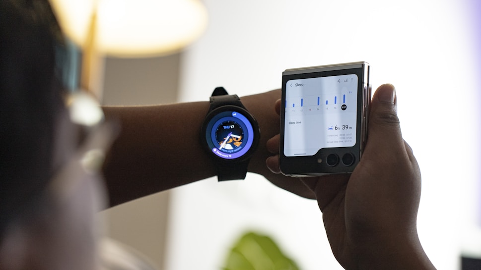 A person is interacting with two electronic devices; a smartwatch on their wrist displaying the time and a cartoon image, and a foldable smartphone showcasing sleep tracking data with graphs and time details. The setting is indoors with soft lighting.