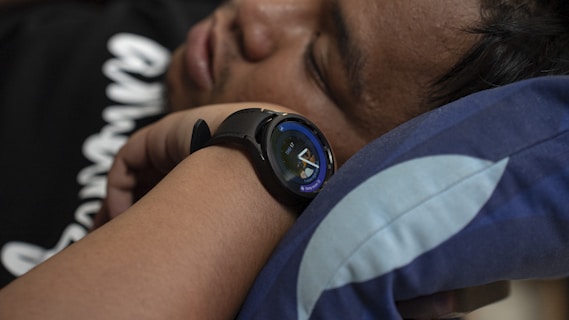 A person is lying down with their head resting on a pillow, wearing a smartwatch on their wrist. The pillow has a dark blue pattern with white accents.