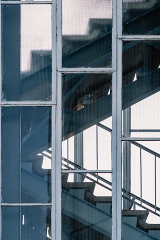 A metal staircase is seen through a grid of glass window panes, with a partial view of the steps and railings behind the textured glass. The image captures the geometric lines and angles of the staircase structure with a backlight illuminating the scene.