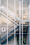 A modern staircase with blue railings and concrete steps is visible through a large grid of windows. The building has a clean, minimalist architecture. A decorative flower and bird mural is present at the bottom of the stairs. There is a prominent sign with the letter 'F' on one of the window panes.