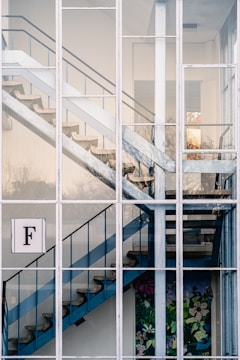 A modern staircase with blue railings and concrete steps is visible through a large grid of windows. The building has a clean, minimalist architecture. A decorative flower and bird mural is present at the bottom of the stairs. There is a prominent sign with the letter 'F' on one of the window panes.