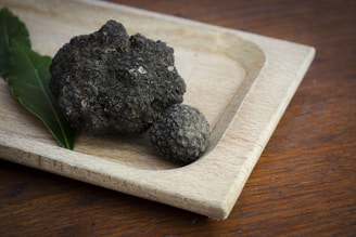 A wooden tray holds two black truffles and a green leaf resting on its light brown surface. The truffles have a rough texture and are positioned on the left side of the tray, while the tray itself is placed on a darker wooden table.