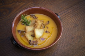 A rustic ceramic bowl filled with creamy orange soup garnished with croutons, crispy bacon bits, and a sprig of rosemary. The bowl is placed on a wooden surface, adding to the warm and cozy ambiance.