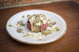 A dish served on a rustic white plate features a stack of raw meat topped with shavings of truffle and pieces of cheese. Small green leaves and red stems, possibly microgreens, are artfully scattered around the plate. The presentation is elegant and minimalistic, highlighting the gourmet ingredients.
