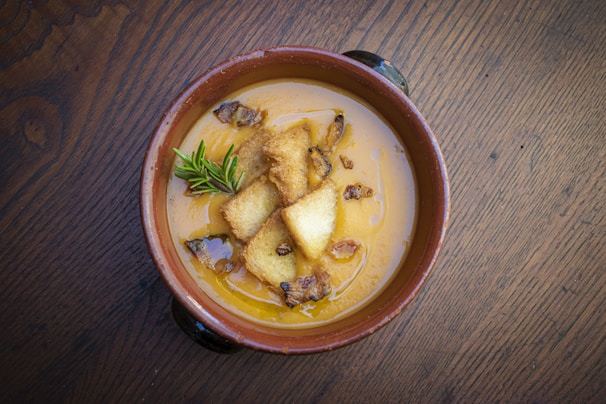 A bowl of creamy orange soup garnished with croutons, crispy bacon bits, and a sprig of fresh rosemary. The soup is served in a rustic brown earthenware bowl on a dark wooden table, adding a warm, cozy feel to the setting.