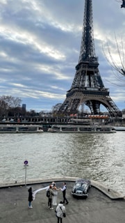 A picturesque scene by the Seine River with the Eiffel Tower towering in the background. A group of people, likely a wedding party, is gathered near a vintage car, with one person holding up a white veil. The cloudy sky adds a dramatic touch to the scene.