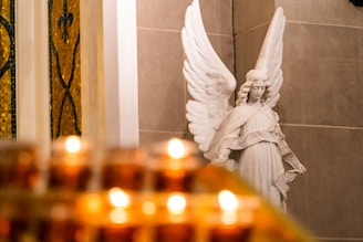 A marble angel statue with extended wings positioned against a stone wall, alongside a stained glass window. The foreground is filled with numerous lit amber candles, casting a warm glow.