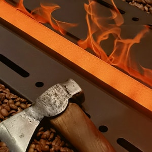 A metal beam glows with intense heat and is surrounded by bright orange flames. In the foreground, a rugged metal and wooden-handled hatchet rests amidst scattered coffee beans, creating a striking contrast between the industrial setting and the organic elements.